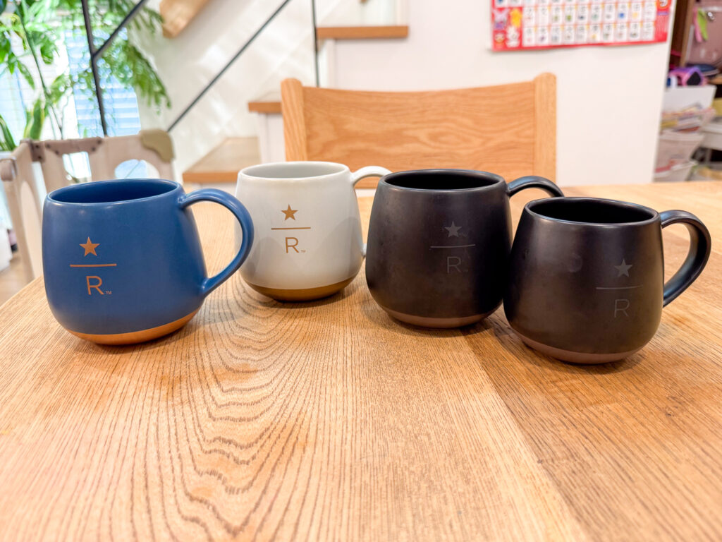 木のテーブルに並べたスターバックス リザーブのマグカップ4つ。左から青、白、黒、黒で、それぞれに星とRのロゴが入っている